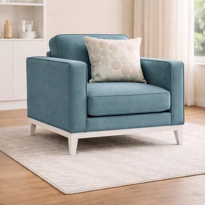 Blue armchair with a patterned pillow on a wooden floor in a room with white walls and a window.
