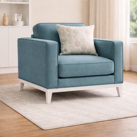Blue armchair with a patterned pillow on a wooden floor in a room with white walls and a window.