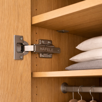 Hafele cabinet hinge on a wooden shelf with pillows and hangers in the background