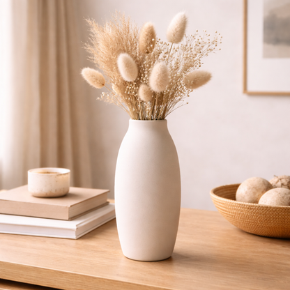 White vase with dried plants on a wooden table, with books and a bowl in the background.